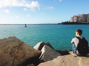 Rear view of man sitting on rocks while looking at sea against sky