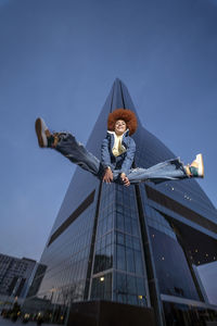 Smiling woman in redhead afro hairstyle jumping in front of skyscraper