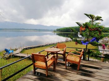 Empty chairs and table by lake against sky