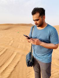 Young man using mobile phone while standing on sand in dessert against sky