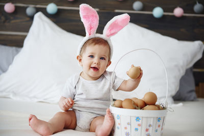 Portrait of cute baby girl with eggs sitting on bed at home
