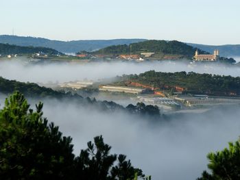 Buildings and trees covered with clouds against clear sky on sunny day