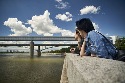Women leaning on railing by river against sky