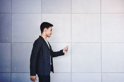 Side view of young businessman using mobile phone against wall