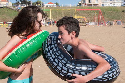 Side view of siblings with inflatable rings standing at beach during sunny day