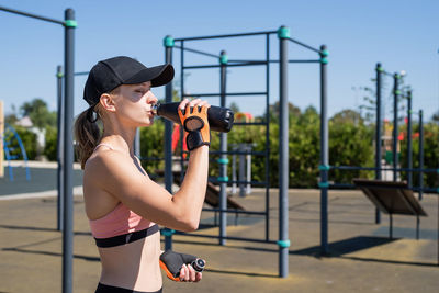 Young sportive woman in sport gloves drinking water from thermos bottle on sport ground