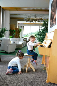 Side view of girl sitting on carpet by sister playing piano at home