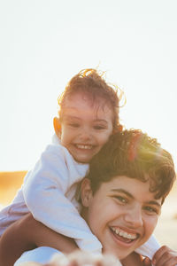 Portrait of happy boy carrying sister on shoulders at beach against clear sky during sunset