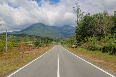 Empty road along trees and mountains against sky