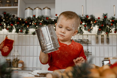 A little boy with iron sieve and dirty from flour helps to cook christmas ginger biscuit in kitchen.