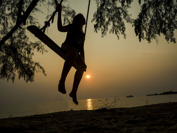 Silhouette girl swinging at beach against sky during sunset