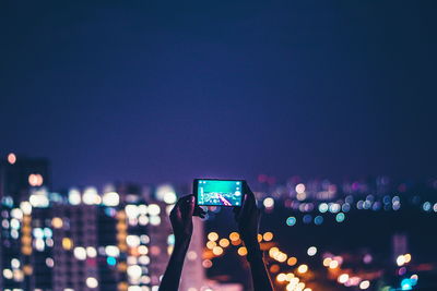 Cropped image of hand photographing illuminated cityscape against sky at night