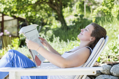 Side view of smiling woman with newspaper relaxing on lounge chair at yard