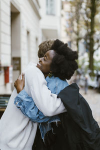 Side view of happy woman embracing boyfriend with arm around while standing on sidewalk in city
