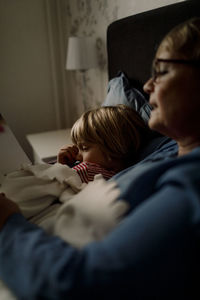 Senior woman reading story book for grandson on bed at home