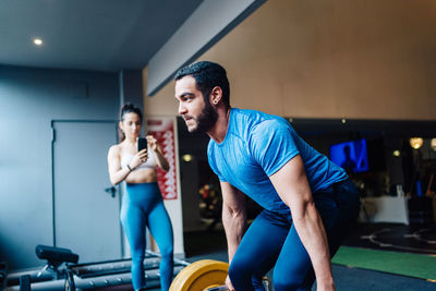 Side view of couple at gym. girl is taking a video on her phone of her partner while lifting