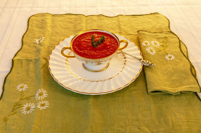 Tomato soup in a white and gold bowl on an embroidered green placemat.