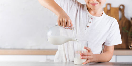 Cute smiling boy pours fresh milk from a jug into a mug in the kitchen.