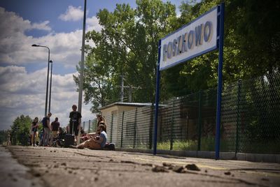 People sitting on road by trees in city against sky