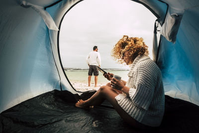 Woman using smart phone in tent while man looking at sea