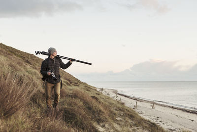 Photographer looking away while carrying tripod on hill by sea