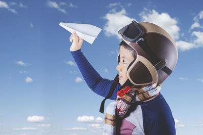 Side view of boy holding paper airplane while wearing helmet against cloudy sky