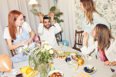 Playful man holding pineapples while sitting amidst female friends at home during dinner party