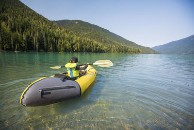 Rear view of young boy paddling yellow boat on scenic lake.