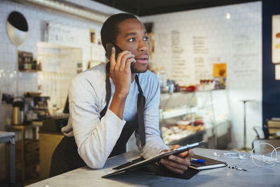 Confident young male employee talking through smart phone while leaning at delicatessen