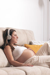 Young pregnant woman listening to music with headphones while relaxing on the couch at home