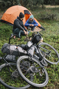 Cyclists arranging halt with warm clothes resting on camp having warm beverages at green meadow at night