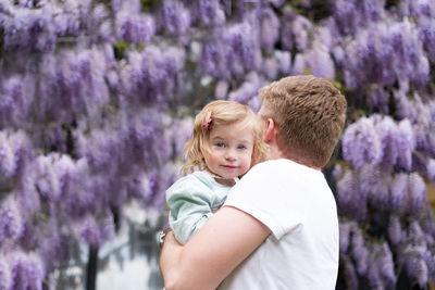 Blond man holding cute baby girl,daughter,looking at camera,near glucinum flowers,purple tree