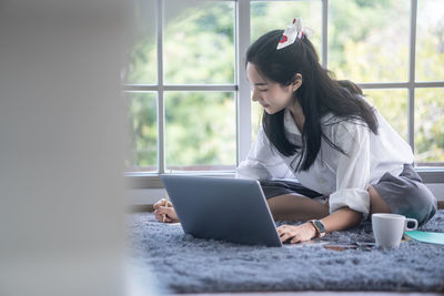 Young woman using mobile phone while sitting on window