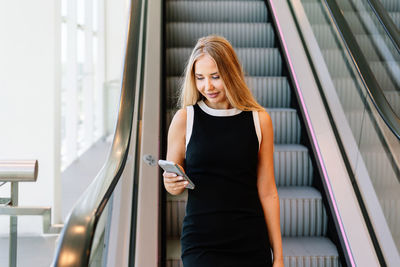 Blond female in black dress using smartphone and looking at screen with smile while standing near moving stairs in modern mall