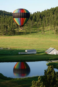 Colorful hot air balloon flying over lake mirror with reflection against sky and pine trees