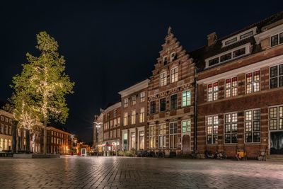 Illuminated buildings by wet street against sky at night