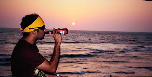 Side view of man drinking beer at beach during sunset