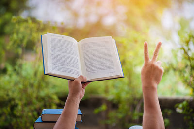 Close-up of person holding book while showing peace sign outdoors