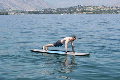 Shirtless man exercising on paddleboard in sea against mountains