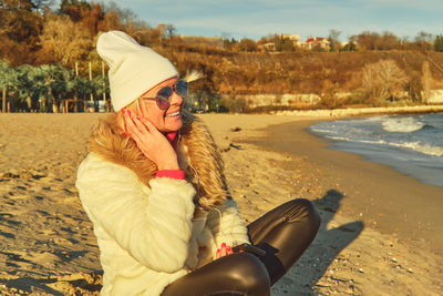 A woman in a fur coat and hat sits on a sandy beach and listens to music on a smartphone.
