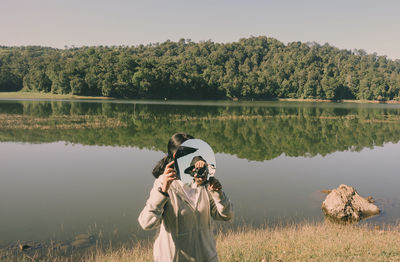 Woman covering face with mirror against lake in sunny day