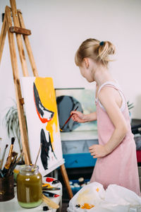 A little cute girl paints a big picture with acrylic at home on an easel.