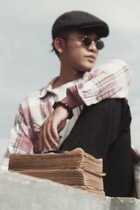 Low angle view of young man with books wearing sunglasses while sitting on retaining wall against sky