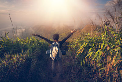 Rear view of mid adult woman with arms outstretched standing amidst plants on mountain against cloudy sky