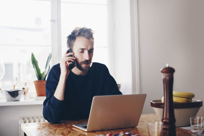 Man talking on mobile phone using laptop at home