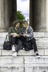 Full length of female friends using mobile phone while sitting on steps