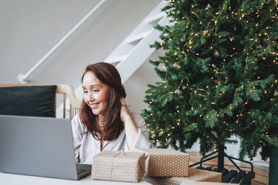 Young adult forty years woman with present gift box using laptop in living room with christmas tree 