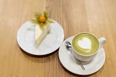On the wooden surface stands a white cup of matcha tea. on a saucer is a white cheesecake with 