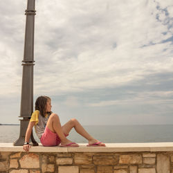 Girl sitting on bridge over sea against sky