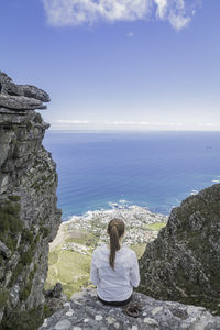 Rear view of woman sitting on rock by sea against sky